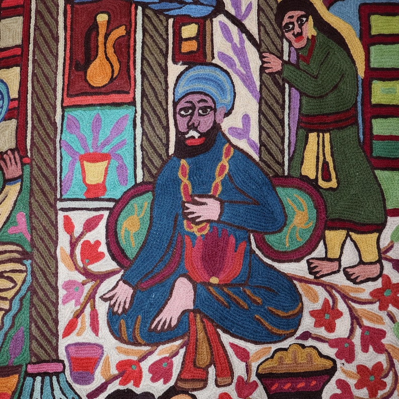 May include: A colorful embroidered artwork depicting a man wearing a blue turban and a blue robe sitting on a cushion. The man is holding a beaded necklace and has a beard. The artwork is set against a background of a building with windows and a floral design.