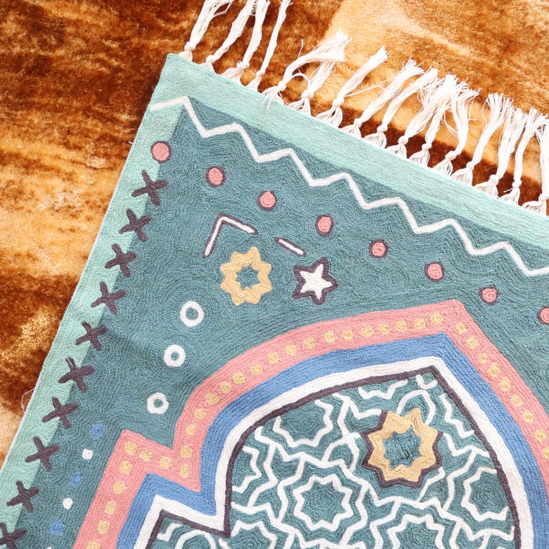 May include: A teal blue rug with a geometric pattern in pink, yellow, and white. The rug has a fringe border.