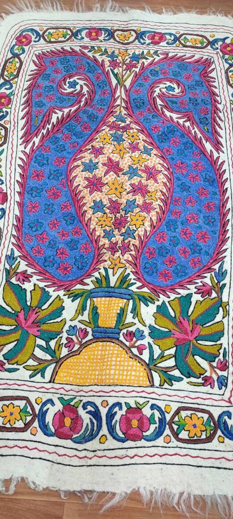 May include: A white fabric rug with a colorful embroidered design. The design features a vase with flowers and leaves, all in shades of pink, blue, yellow, and green.