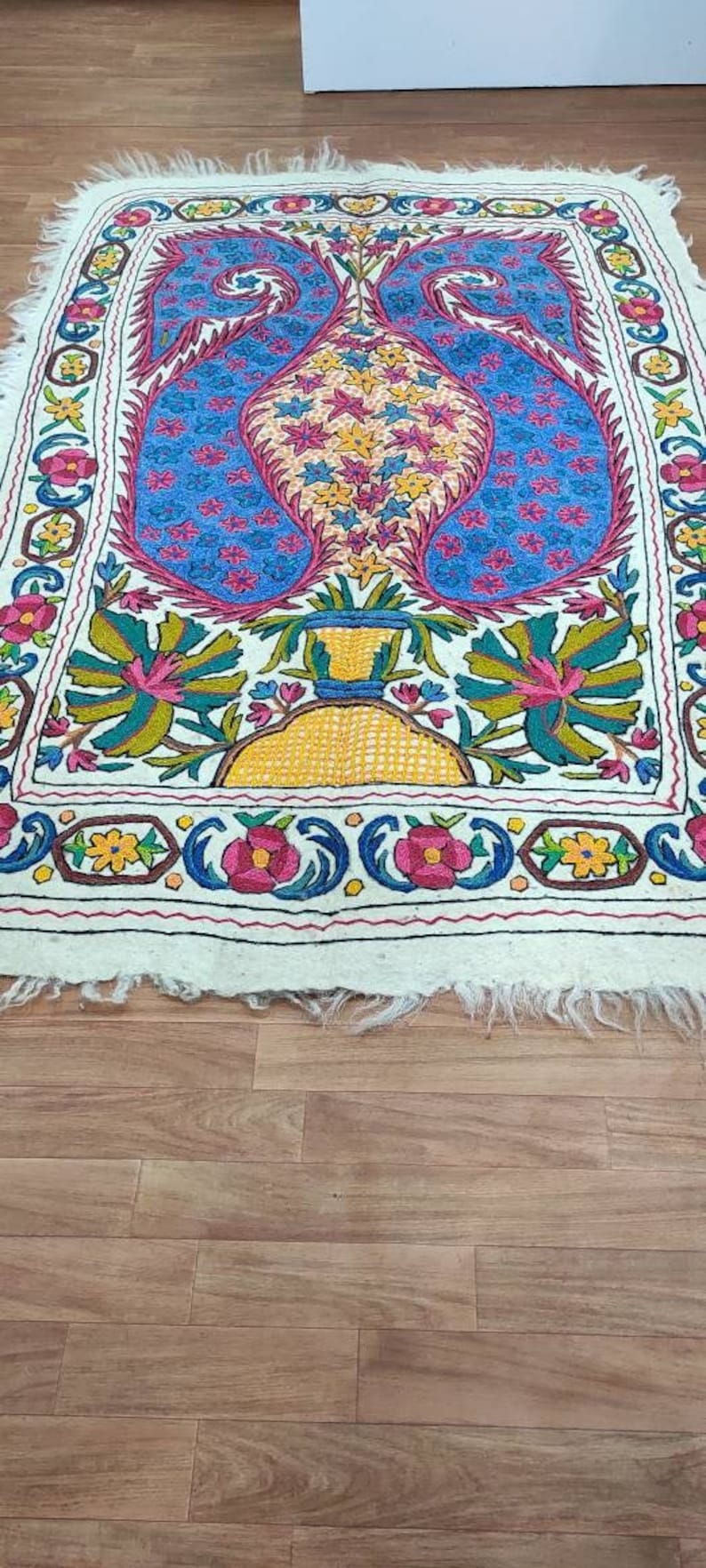 May include: A white embroidered rug with a floral design featuring a vase with two peacocks. The rug has a border of flowers and leaves in shades of pink, yellow, blue, and green.