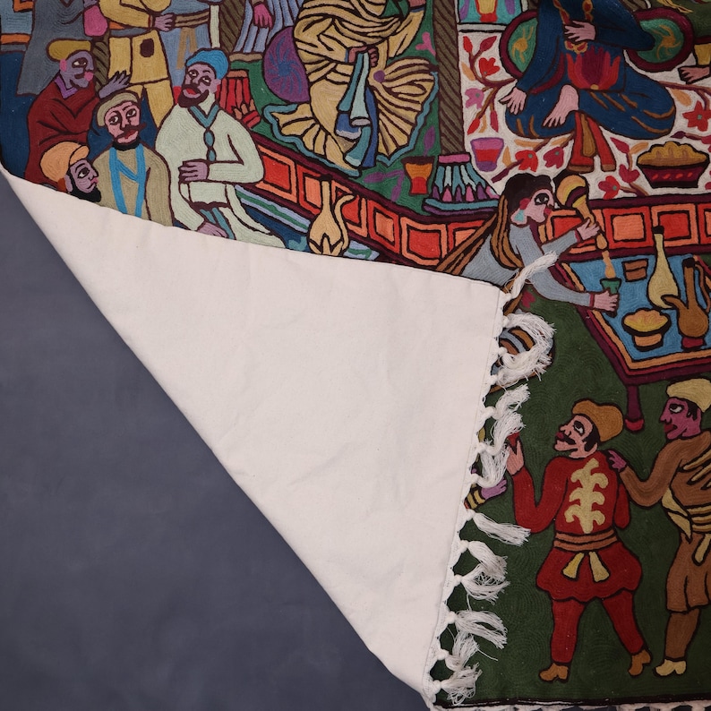 May include: A colorful tapestry with a green background depicting a scene of people gathered around a table. The tapestry has a white fringe border and is folded over on itself.