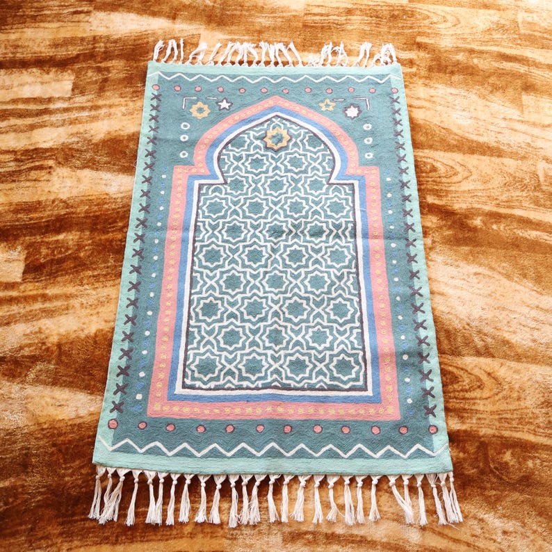 May include: A teal blue prayer rug with a geometric pattern and a pink and yellow border. The rug has a white fringe around the edges.