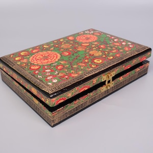 Może przedstawiać: Czarne drewniane pudełko z pokrywką na zawiasach. Pudełko jest ozdobione wzorem kwiatowym w odcieniach czerwieni, zieleni i złota. Pudełko ma mosiężny zamek.