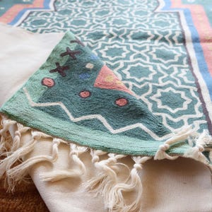 May include: A teal green prayer rug with a geometric pattern and white fringe. The rug has a pink and blue design in the center.