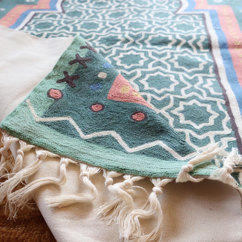 May include: A teal green prayer rug with a geometric pattern and white fringe. The rug has a pink and blue design in the center.