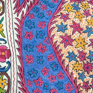 May include: A white fabric with intricate embroidery featuring a blue and pink paisley design with floral motifs. The paisley is surrounded by smaller flowers in shades of pink, yellow, and blue.
