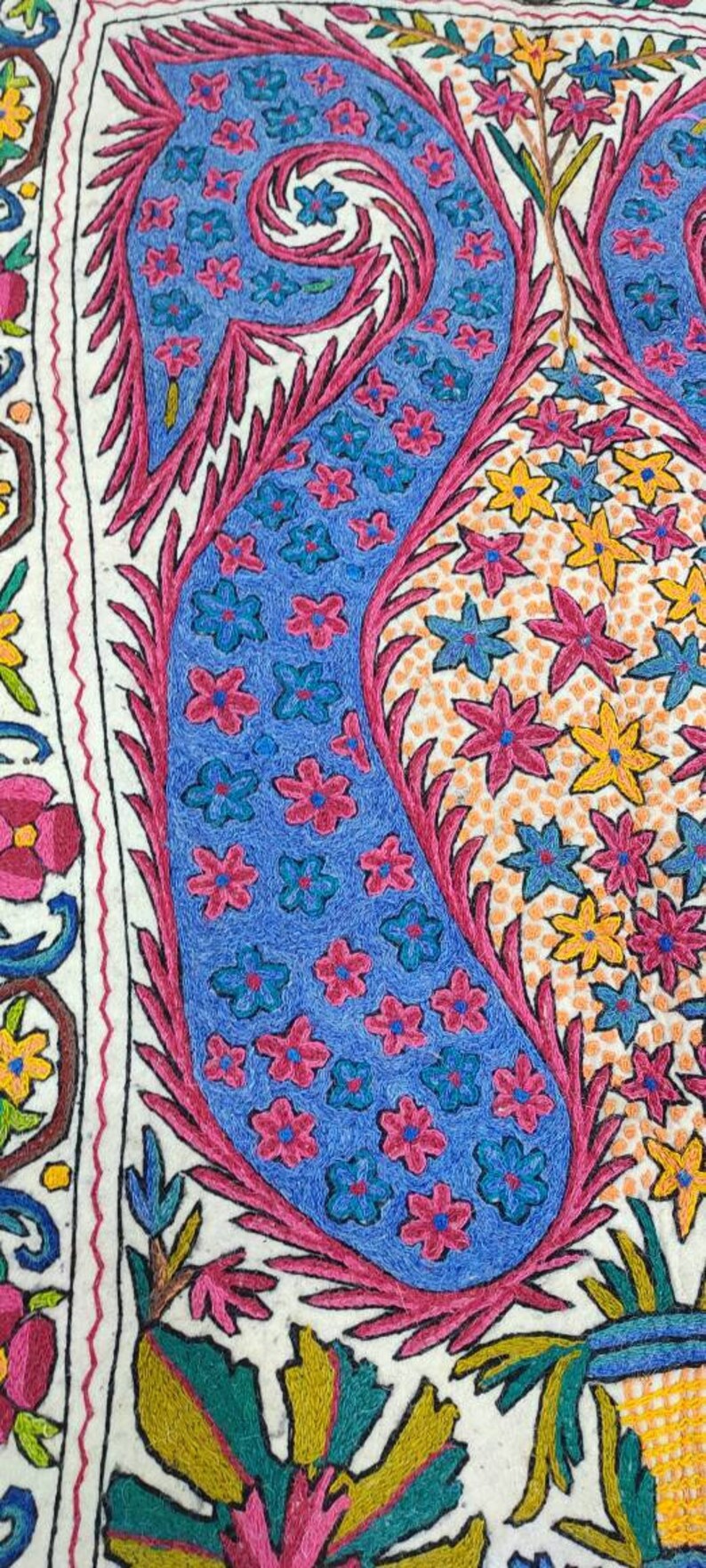 May include: A white fabric with intricate embroidery featuring a blue and pink paisley design with floral motifs. The paisley is surrounded by smaller flowers in shades of pink, yellow, and blue.