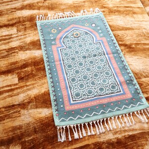 May include: A blue and pink prayer rug with a geometric pattern and white fringe. The rug has a traditional arch design in the center.