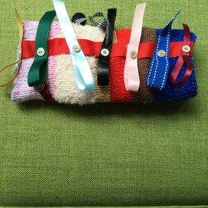 Puede incluir: Un juguete sensorial tejido a mano, colorido, con varias cintas y botones adjuntos. El juguete presenta una mezcla de texturas y colores, incluyendo rosa, verde, azul, rojo y blanco. Las cintas están aseguradas con botones.