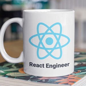 May include: White ceramic mug with a light blue atom symbol and the words "React Engineer" printed on the side. The mug sits on a book with a floral design, placed on a wooden surface.