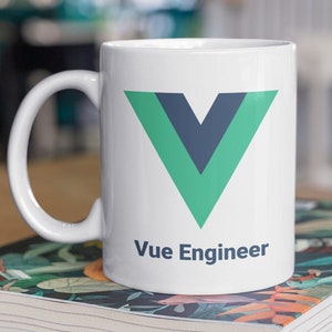 Pode incluir: Caneca de cerâmica branca com o logotipo Vue.js, um V verde e azul escuro, e o texto "Vue Engineer". A caneca está em cima de um livro com um design floral. O fundo está desfocado.