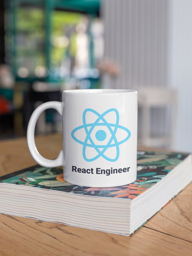 May include: White ceramic mug featuring a light blue atom symbol and the text "React Engineer". The mug is placed on a book with a floral pattern, resting on a wooden table.