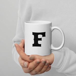 Può includere: Una tazza di ceramica bianca con la lettera "F" nera stampata sopra. La tazza ha un manico curvo e viene tenuta con entrambe le mani. Il design è semplice e audace, rendendola un pezzo d'effetto.
