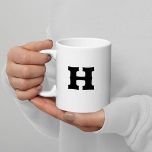 Puede incluir: Taza de cerámica blanca con la letra negra "H". La taza tiene un asa curvada y una superficie lisa y brillante. Un diseño sencillo para té, café o cualquier bebida caliente.