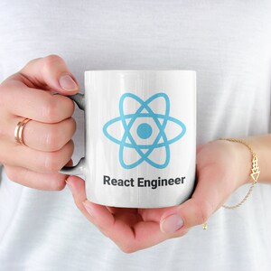 May include: White ceramic mug featuring a light blue atom symbol and the text "React Engineer" in black. The mug is held by a person wearing a white top and gold jewellery.
