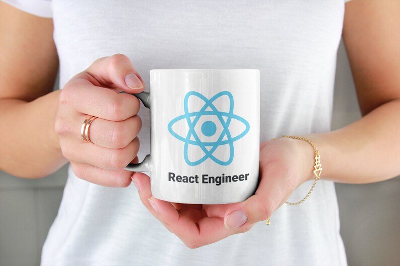 May include: White ceramic mug featuring a light blue atom symbol and the text "React Engineer" in black. The mug is held by a person wearing a white top and gold jewellery.