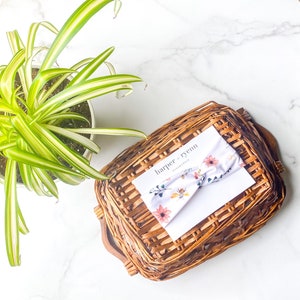 May include: A white headband with a floral pattern and bees. The headband is in a wicker basket with a white card that says "harper + ryenn collection".