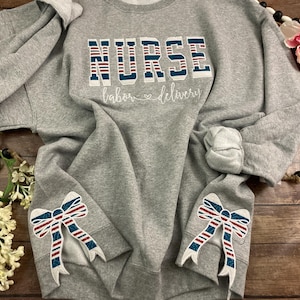 May include: A grey sweatshirt featuring the word "NURSE" in a bold, patriotic font, with "labor & delivery" in script below. The sleeves have bow accents with red, white, and blue stripes. The sweatshirt is displayed on a wooden surface.
