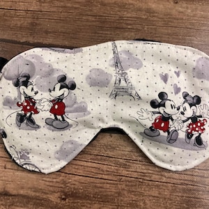 May include: Sleep mask featuring Mickey and Minnie Mouse in Paris. The mask has a white background with gray polka dots, clouds, and the Eiffel Tower. Mickey and Minnie are depicted in red and black outfits. Black ear-shaped accents are on each side.