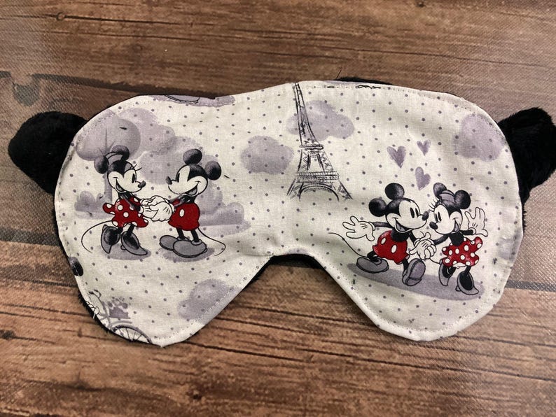 May include: Sleep mask featuring Mickey and Minnie Mouse in Paris. The mask has a white background with gray polka dots, clouds, and the Eiffel Tower. Mickey and Minnie are depicted in red and black outfits. Black ear-shaped accents are on each side.
