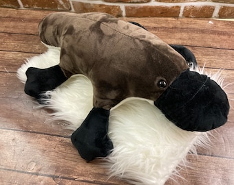 May include: A brown and black plush platypus toy lying on a white fluffy rug. The platypus has a long, narrow snout and webbed feet.