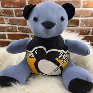 May include: A handmade blue teddy bear with black accents on the ears and nose. The bear's chest features a black, yellow, and white graphic design. The bear is sitting on a fluffy white surface.