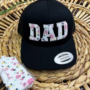 May include: Black mesh trucker hat with a white floral embroidered "DAD" on the front. The hat has a black adjustable strap and a white label that says "The Authentic Snapback Yupoong Since 1974".