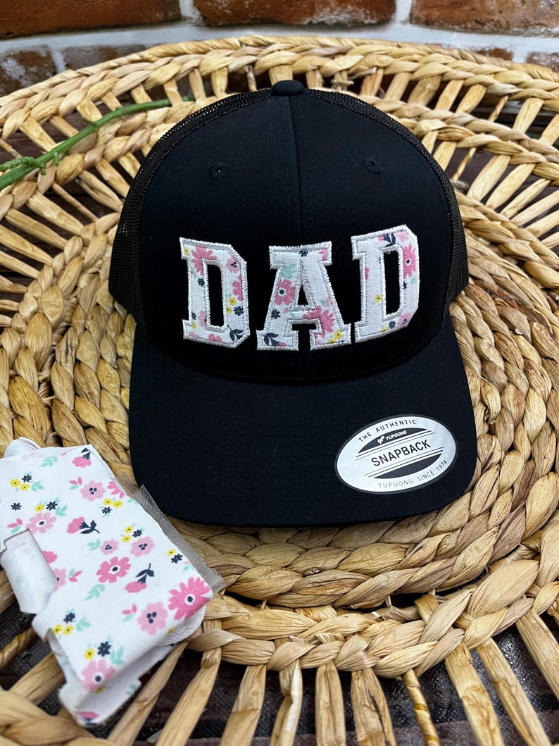 May include: Black mesh trucker hat with a white floral embroidered "DAD" on the front. The hat has a black adjustable strap and a white label that says "The Authentic Snapback Yupoong Since 1974".