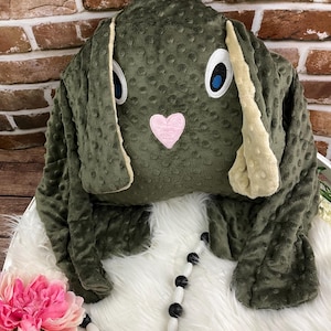 May include: A green plush bunny-shaped weighted blanket with a pink heart nose and blue eyes. The blanket is lying on a white fluffy surface with black and white beads.