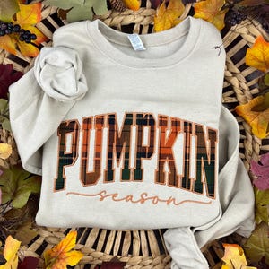 May include: A beige crewneck sweatshirt featuring the word PUMPKIN in a large, orange, green, and black plaid block font, with the word season written in a smaller, cursive orange script font centred below the plaid lettering.