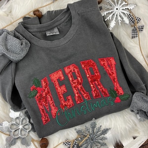 May include: A dark gray sweatshirt with the words "MERRY Christmas" in red sequined letters and green script. The design includes holly leaves and berries. The sweatshirt is displayed on a white fluffy surface with decorative snowflakes.