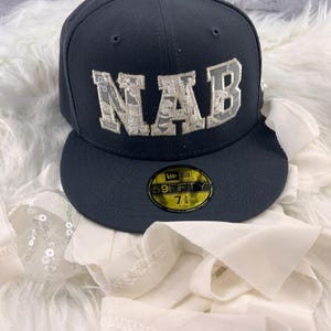 May include: A dark grey New Era 59FIFTY fitted cap featuring the letters NAB in white lace-patterned applique on the front. A circular gold and black sticker on the flat brim displays the text 59FIFTY and the size 7 3/8.