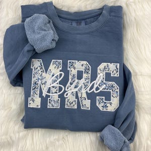 May include: A blue sweatshirt with the word "MRS" in large, floral-patterned letters and the name "Byrd" in cursive script. The sweatshirt has a crew neck and long sleeves with ribbed cuffs. The garment is displayed on a white, fluffy surface.