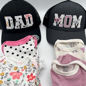 May include: Two black and grey mesh trucker hats with the words "DAD" and "MOM" embroidered in colorful fabric. Four baby onesies in pink, white, and black with polka dots, floral, and striped patterns.