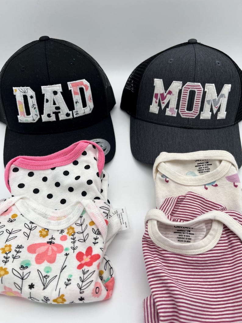 May include: Two black and grey mesh trucker hats with the words "DAD" and "MOM" embroidered in colorful fabric. Four baby onesies in pink, white, and black with polka dots, floral, and striped patterns.