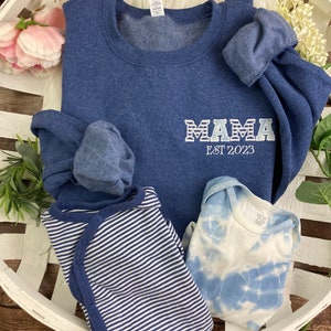 May include: A blue sweatshirt with the text "MAMA EST 2023" in white.  A blue and white striped baby onesie and a blue and white tie-dye baby onesie are also in the image.