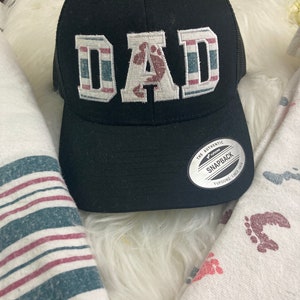 May include: Black and white baseball cap with a red, white, and blue striped design. The word "DAD" is embroidered on the front of the cap in white with a red footprint in the center of the "A". The cap has a mesh back and a black adjustable strap.