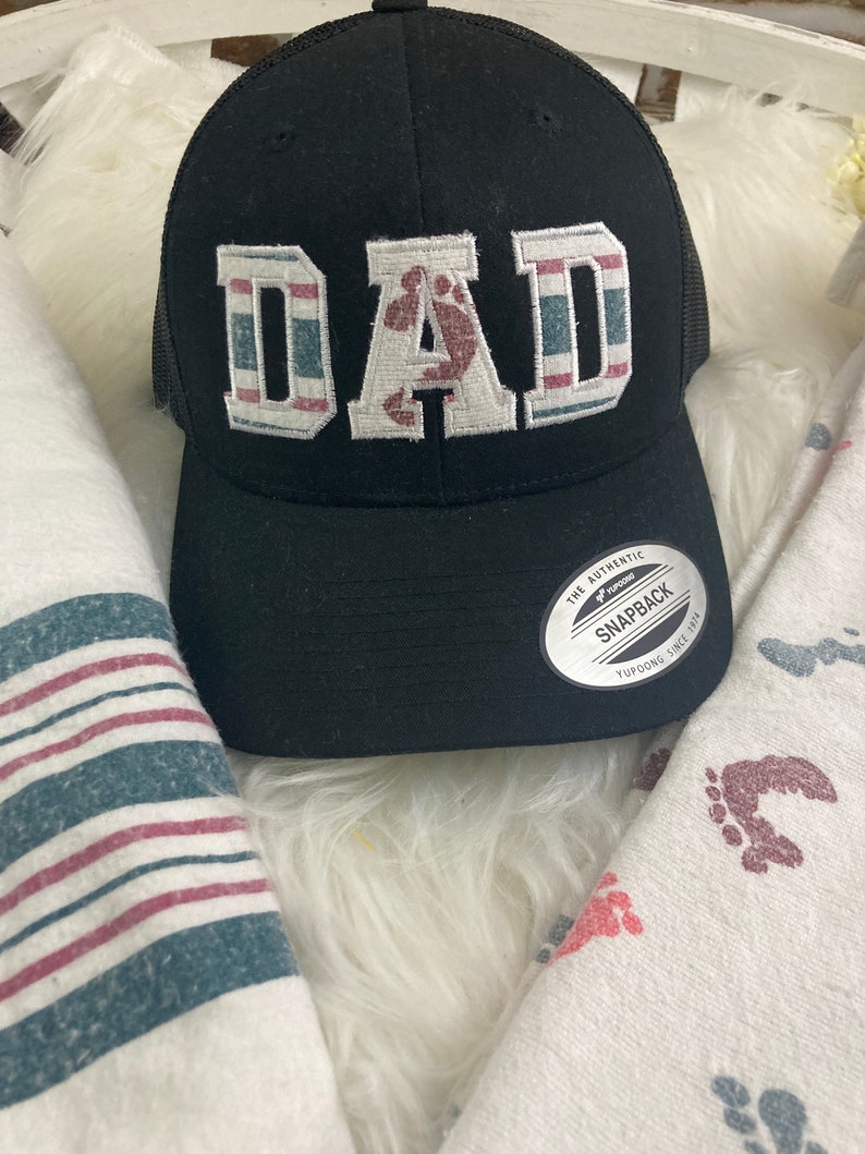 May include: Black and white baseball cap with a red, white, and blue striped design. The word "DAD" is embroidered on the front of the cap in white with a red footprint in the center of the "A". The cap has a mesh back and a black adjustable strap.