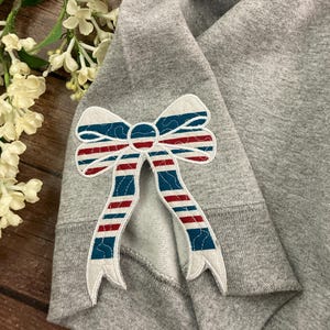 May include: Gray sweatshirt with a red, white, and blue striped bow applique on the sleeve.