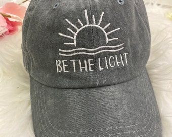 May include: A gray baseball cap with a curved brim. The cap features white embroidered text and a sun design. The text reads "BE THE LIGHT". The cap is made of a soft, textured material.