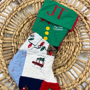 May include: A quilted Christmas stocking with a green elf-themed top, featuring the initials "JT" in red. The stocking has various patches with Christmas motifs, including candy canes, a car carrying a tree, and the text "My First Christmas."