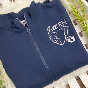 May include: A navy blue zip-up sweatshirt with a white embroidered design. The design includes the text "Jill RN" with a heart and a stethoscope, a baby sleeping inside the heart, and two footprints.