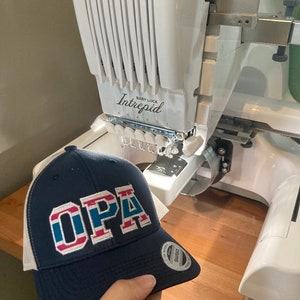 May include: A blue and white trucker hat with the word "OPA" embroidered in red, white, and blue. The hat is being held in front of a white embroidery machine with the brand name "Baby Lock Intrepid" printed on it.