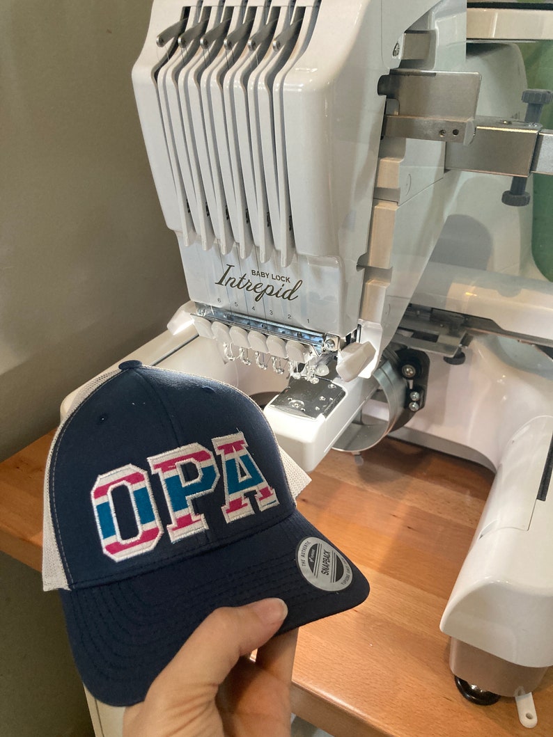 May include: A blue and white trucker hat with the word "OPA" embroidered in red, white, and blue. The hat is being held in front of a white embroidery machine with the brand name "Baby Lock Intrepid" printed on it.