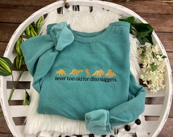 May include: A teal crewneck sweatshirt featuring gold embroidered dinosaur silhouettes and the black embroidered text "never too old for dino nuggets" centered on the chest. The garment has long sleeves and a ribbed collar, cuffs, and hem.