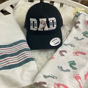 May include: A black and white trucker hat with the word "DAD" embroidered on the front. The hat has a red, white, and blue striped band around the brim. The hat is sitting on a white fuzzy surface.