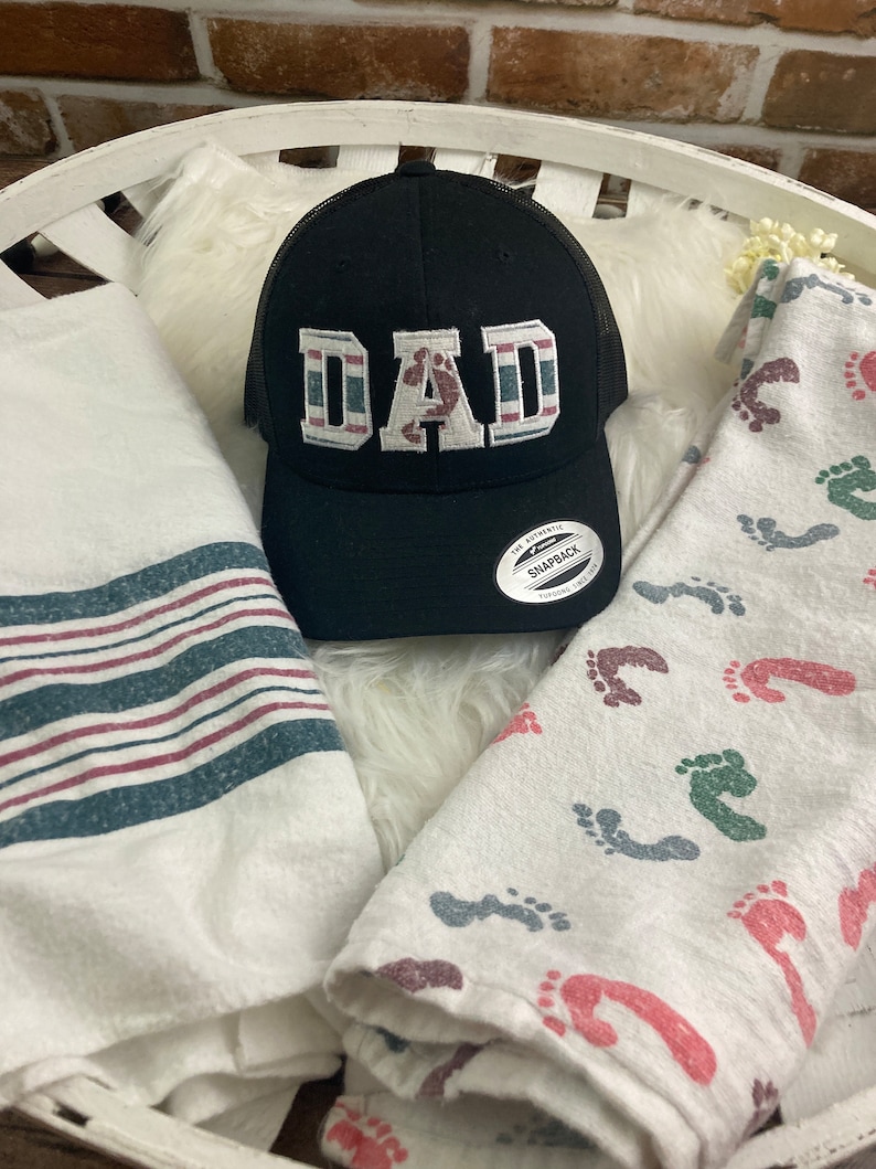 May include: A black and white trucker hat with the word "DAD" embroidered on the front. The hat has a red, white, and blue striped band around the brim. The hat is sitting on a white fuzzy surface.