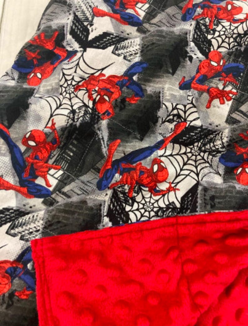 May include: A red and blue Spiderman themed fleece blanket with a red minky backing. The blanket features a repeating pattern of Spiderman characters and webs.