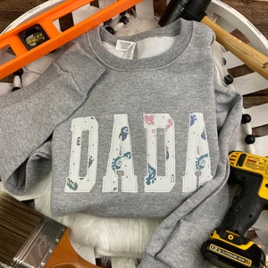 May include: A gray sweatshirt with the word "DADA" in white block letters. The letters are filled with a colorful paw print pattern. The sweatshirt is surrounded by tools, including a level, hammer, and drill, on a white, woven tray.