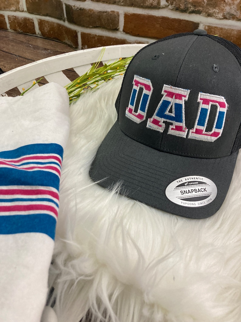 May include: A black and gray trucker hat with a white, blue, and pink "DAD" embroidered on the front. The hat has a mesh back and a white label that reads "The Authentic Yupoong Snapback Yupoong Since 1974".